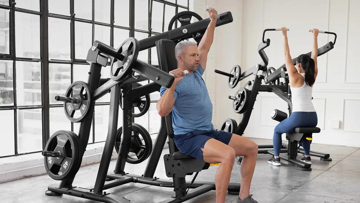 Two people in workout clothes using Matte Black plate-loaded Shoulder Presses in a gym.