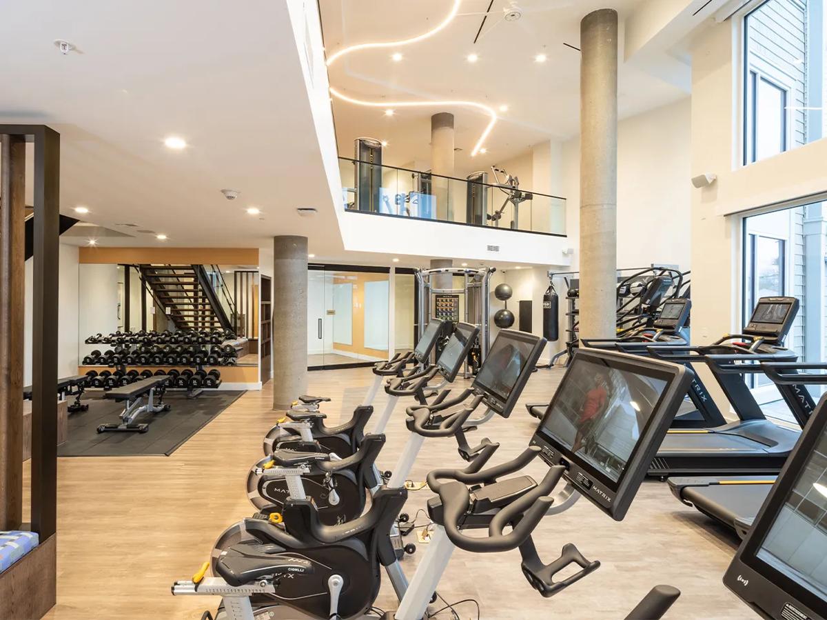 Centre de fitness sur deux niveaux avec appareils d'entraînement cardio, des haltères et de grandes fenêtres laissant entrer la lumière naturelle.