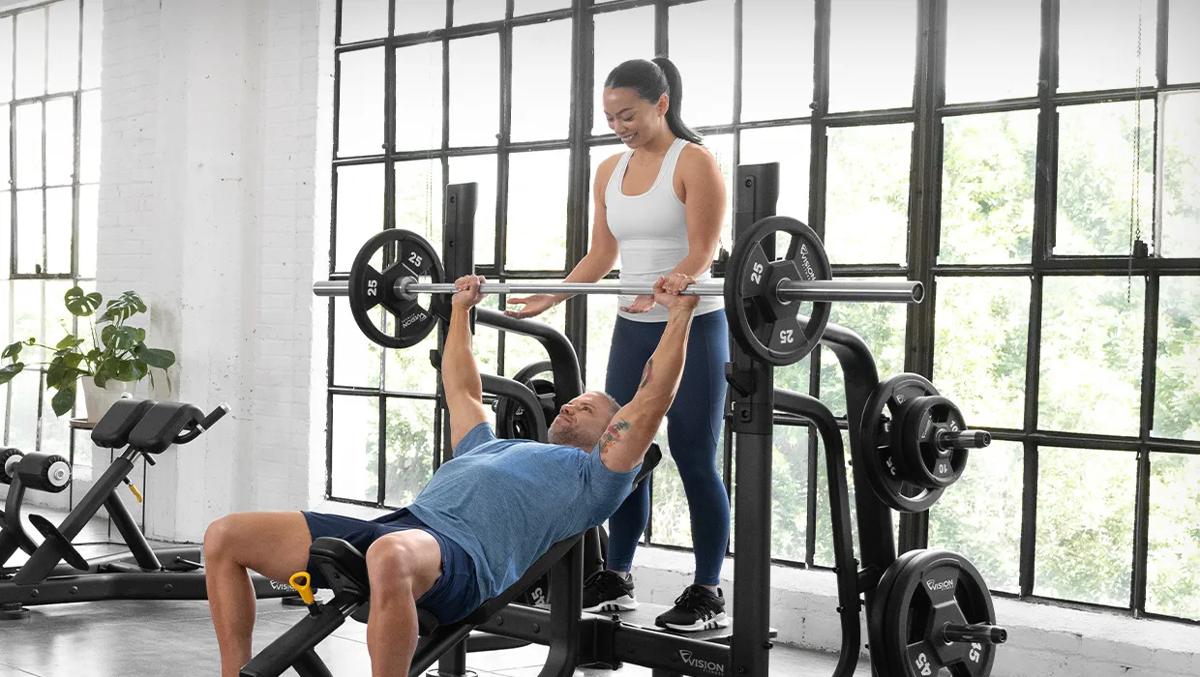 Two people on the Vision Incline Bench assisting while one is lifting with large windows behind them.
