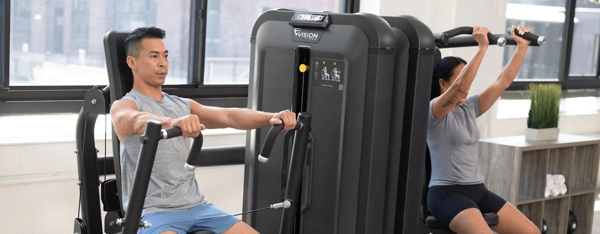 Person using the Vision Fitness Chest Press machine and woman using the Shoulder Press machine, both in Matte Black, exercising side by side in a bright, modern gym.