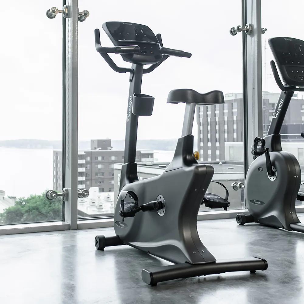 Two Vision Fitness Upright Bikes in a bright gym setting with a cityscape view in the background.