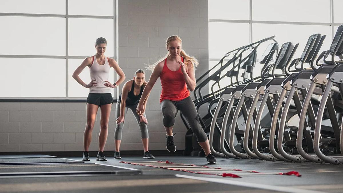 Woman running through a ladder on the gym floor with two other women watching behind her
