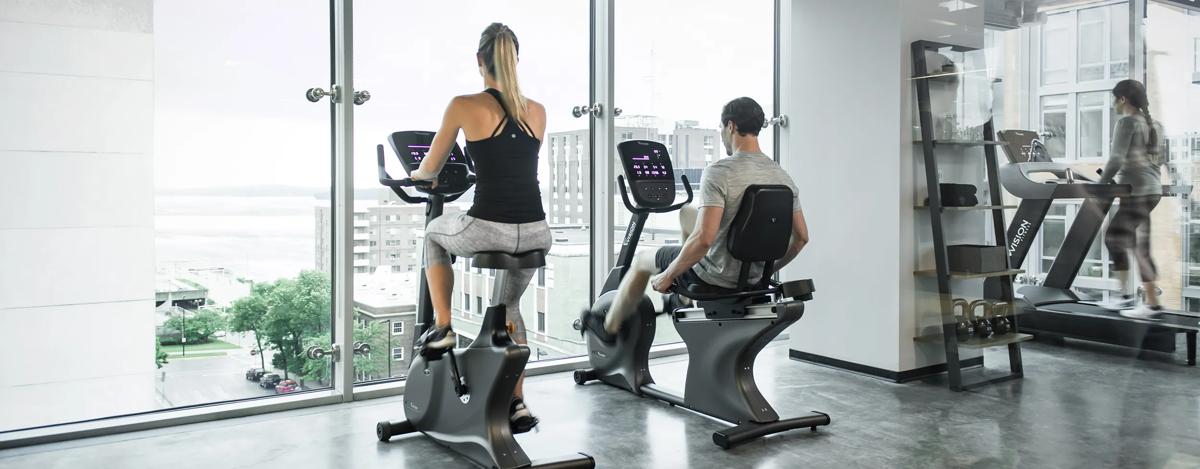 Two people cycling on stationary bikes with digital displays in a modern gym with floor-to-ceiling windows, while another person runs on a treadmill in the background.