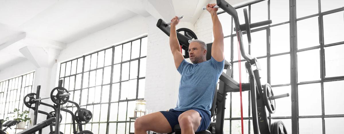 A person in a blue workout shirt doing a shoulder press on the Vision Shoulder Press machine in a bright room with large windows.