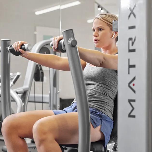 A woman wearing a gray tank top and blue shorts working out on a Matrix strength machine.