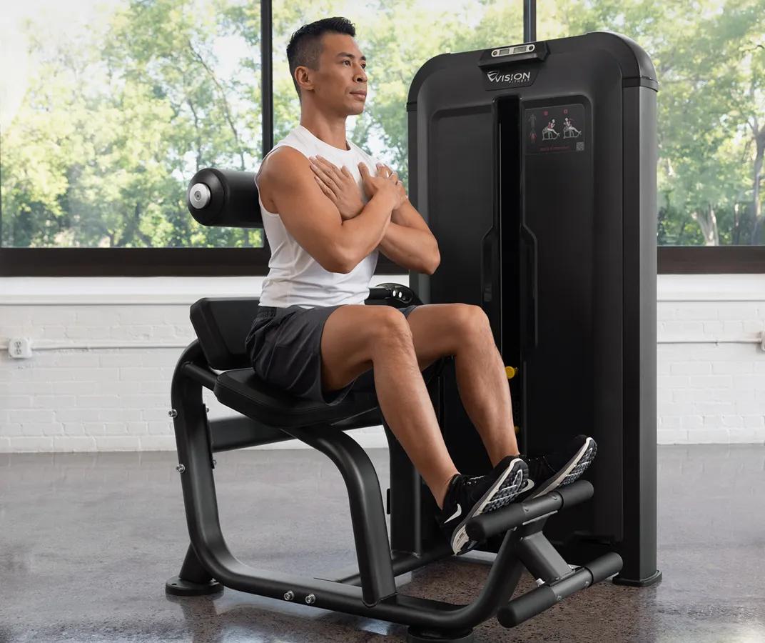 Person seated on the Vision Fitness Back Extension machine with arms crossed, positioned at the start of a back extension exercise in a bright gym.