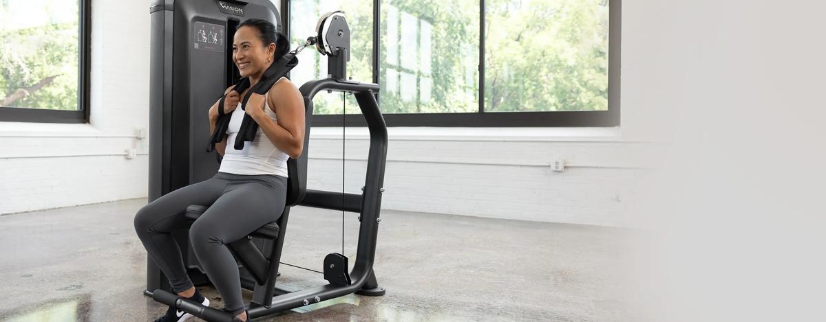 Person seated on the Vision Fitness Abdominal machine with a shoulder harness in place, preparing to perform a crunch in a bright gym setting.