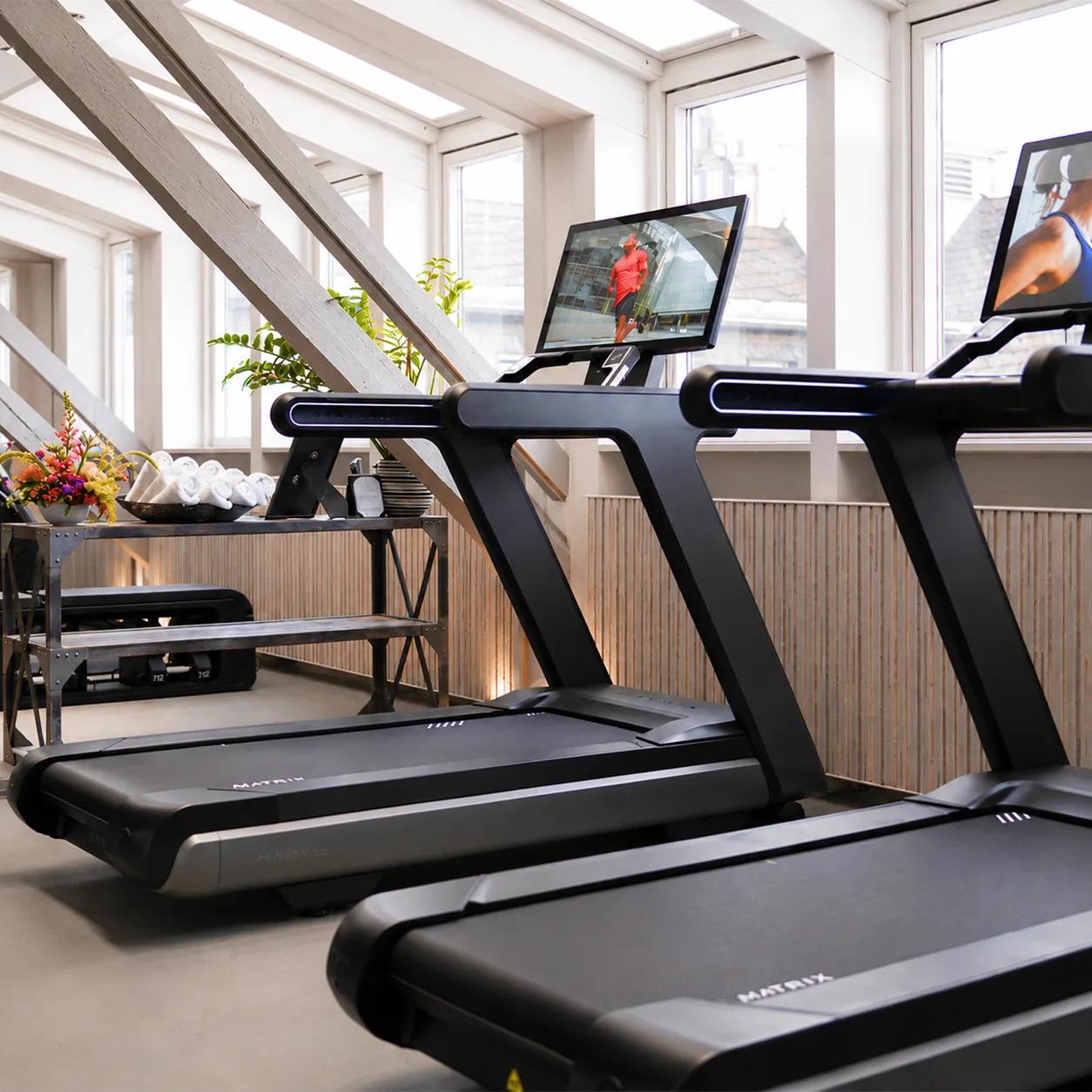 Matzwarte Matrix Fitness-loopbanden in een lichte fitnessruimte met grote ramen en geïntegreerde schermen, naast elkaar opgesteld voor cardiotraining.