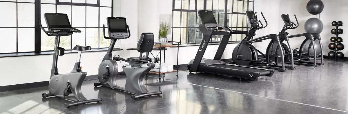 A row of cardio machines, including bikes, treadmills, and ellipticals, in a spacious, well-lit industrial-style gym.