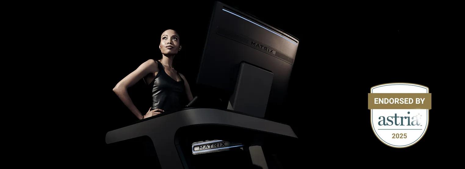 Een persoon staat op een Matrix Fitness-cardiotoestel met een groot scherm in een donkere studio-omgeving, naast een Astria 2025-erkenningsbadge die de erkenning in de sector benadrukt.