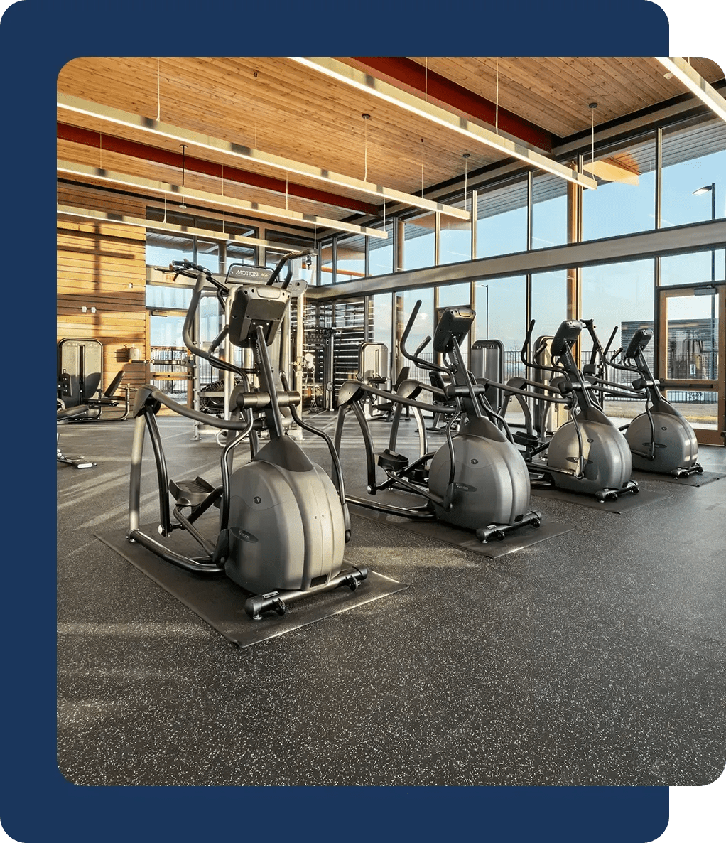 A modern gym with large windows, wooden ceilings, and a row of elliptical machines positioned on a rubberized floor.