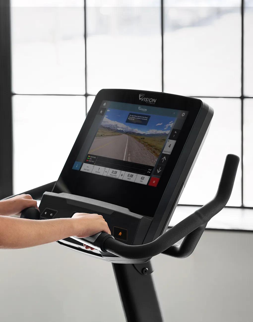 Close-up of a person gripping the handlebars of the Vision Fitness Upright Bike, with a large touchscreen display showing a scenic virtual ride interface.