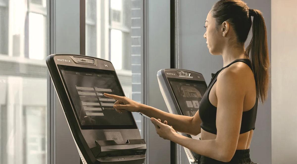 Een persoon gebruikt een Matrix Fitness-touchscreenconsole en tikt op trainingsopties terwijl hij een smartphone vasthoudt in een lichte fitnessruimte met grote ramen.