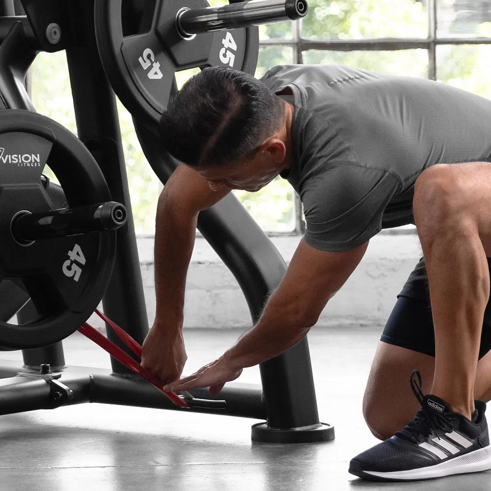 Person in workout clothes attaching red resistance bands to a Matte Black plate-loaded Leg Press in a gym.
