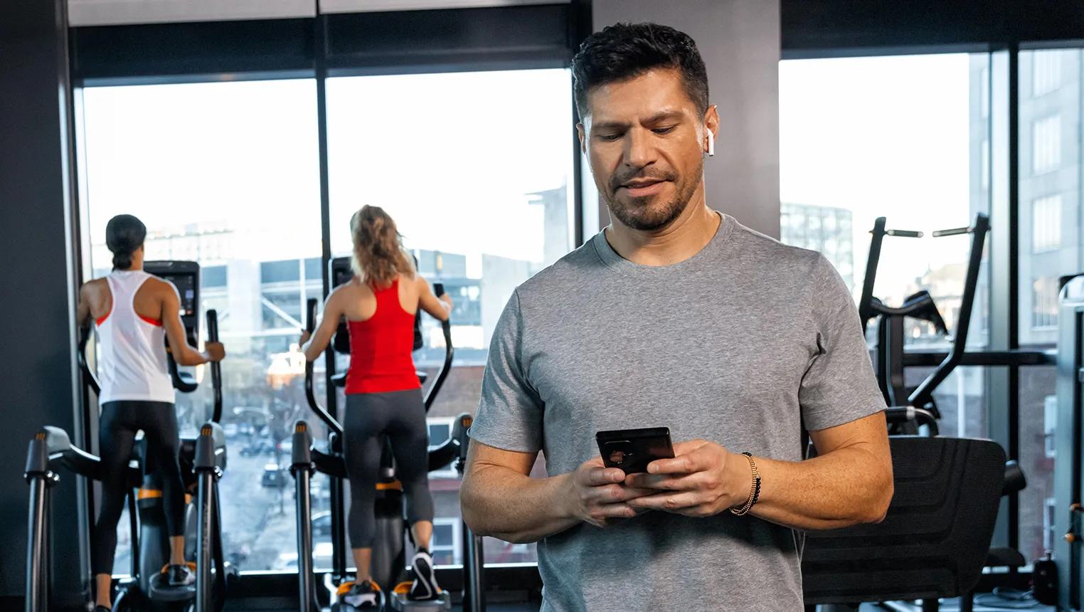 Man in a commercial gym looking at his phone with two women on elliptical machines in the background