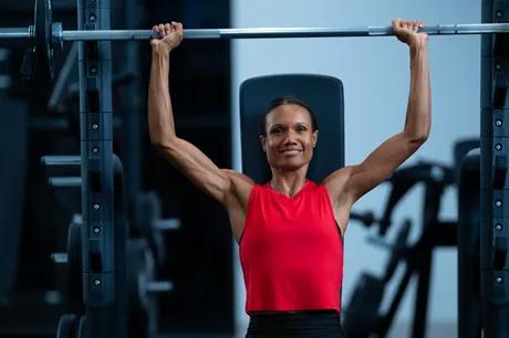 A person in a red workout top sitting on a bench with a loaded barbell facing forward smiling