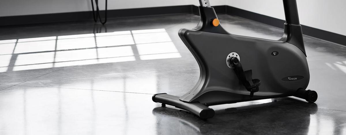 A close-up side view of the Vision Fitness Upright Bike in a gym with a polished concrete floor and large windows reflecting sunlight. The bike's flywheel, pedals, and frame are visible.