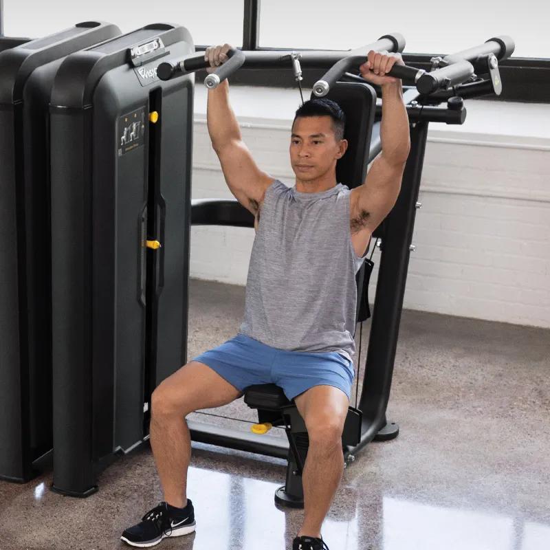 Person using the Vision Fitness Converging Shoulder Press machine in Matte Black, seated with arms bent and handles positioned for an upward pressing motion in a bright gym.