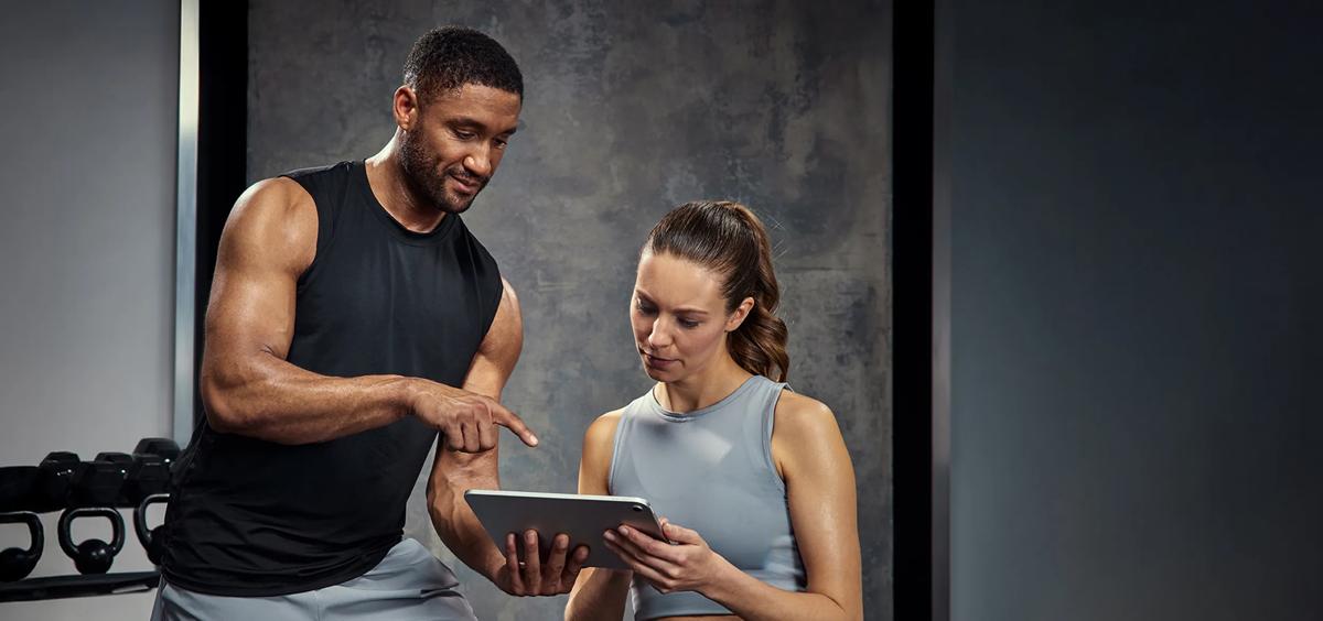Una persona con una maglietta da allenamento nera che indica un tablet grigio mentre un'altra persona con un top da allenamento grigio lo tiene in mano in una palestra.