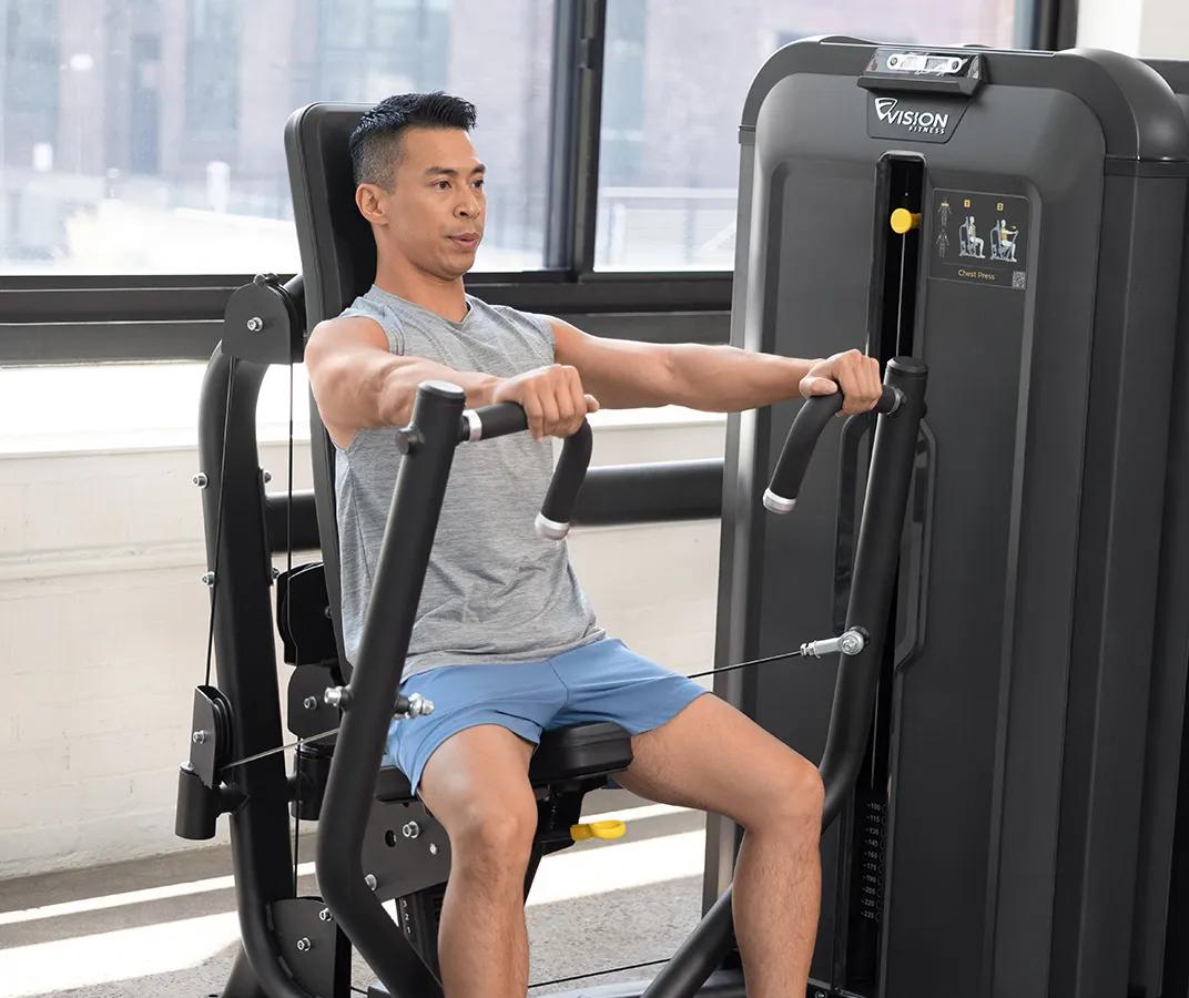 Person using the Vision Fitness Chest Press machine and woman using the Shoulder Press machine, both in Matte Black, exercising side by side in a bright, modern gym.