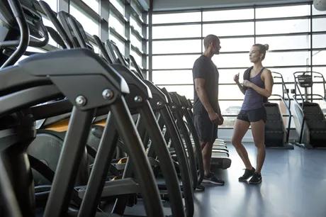 Two people in workout clothing in a gym full of cardio equipment in front of floor to ceiling windows