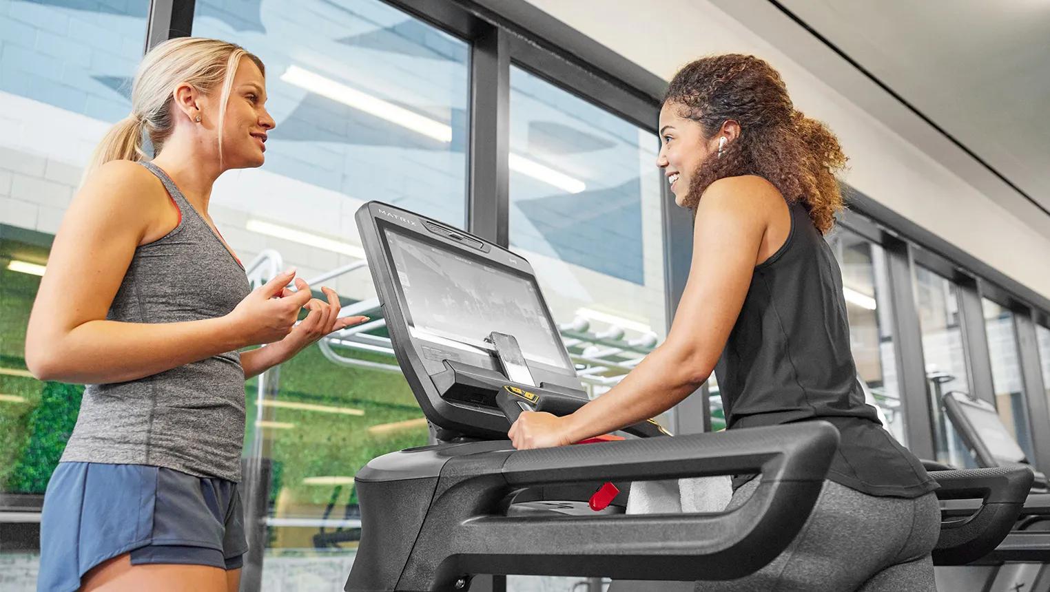 Woman walking on a treadmill at the gym while talking to another woman standing next to her