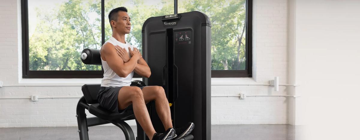 Person seated on the Vision Fitness Back Extension machine with arms crossed, positioned at the start of a back extension exercise in a bright gym.