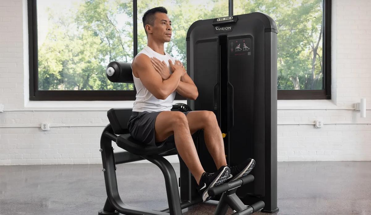 Person seated on the Vision Fitness Back Extension machine with arms crossed, positioned at the start of a back extension exercise in a bright gym.