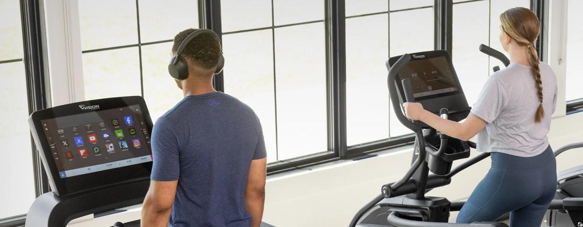 Two people exercising on cardio equipment in a gym setting, the person on the left is using wireless headphones connected to the ENT Console on the treadmill.