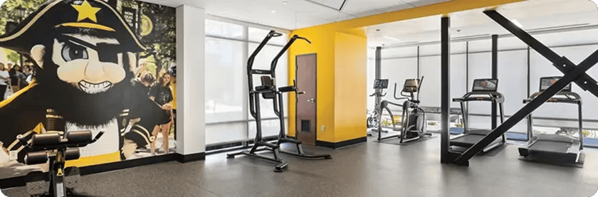 A handful of cardio equipment in a small college gym with a yellow wall