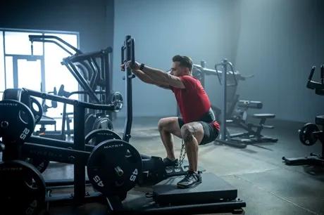 A person in a red tee shirt in a dark gym squatting on an equipment while holding the handells