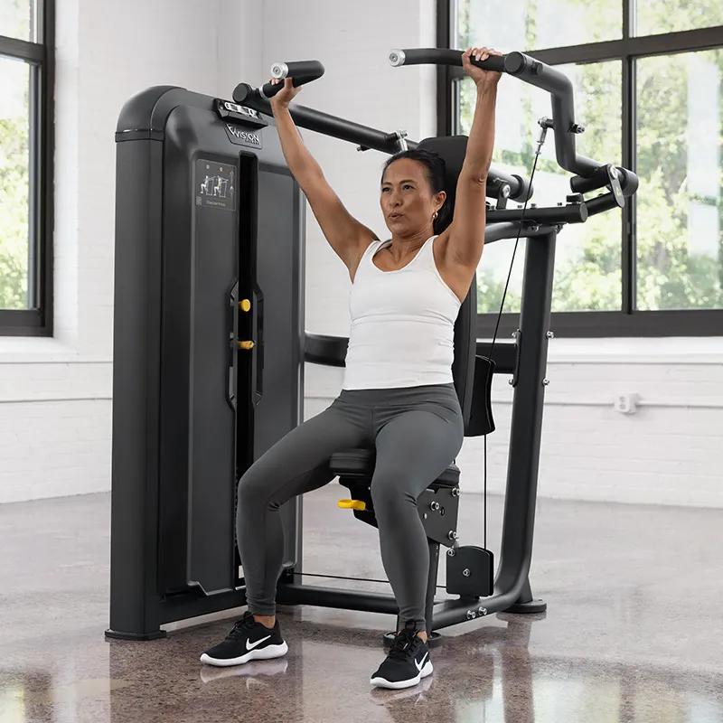 Person completing a shoulder press on the Vision Fitness Shoulder Press machine in Matte Black, arms fully extended overhead while seated in a bright gym.