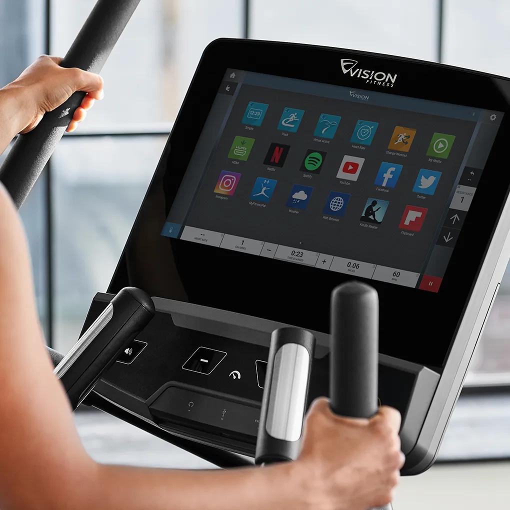 Two people exercising in a well-lit gym, one running on a Vision treadmill with an interactive display and the other using a Vision elliptical trainer.