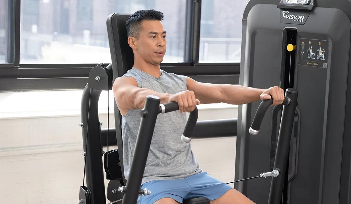 Person using the Vision Fitness Chest Press machine and woman using the Shoulder Press machine, both in Matte Black, exercising side by side in a bright, modern gym.