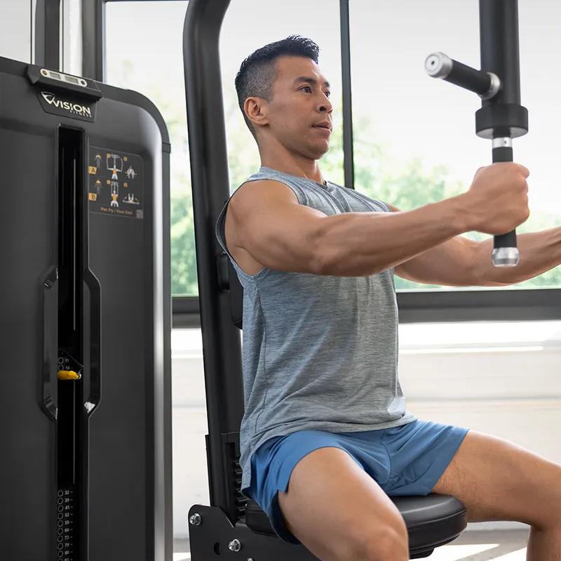 Two people using Vision Fitness strength machines in a bright gym, with the woman in front performing a pec fly exercise on the Pec Fly/Rear Delt machine.