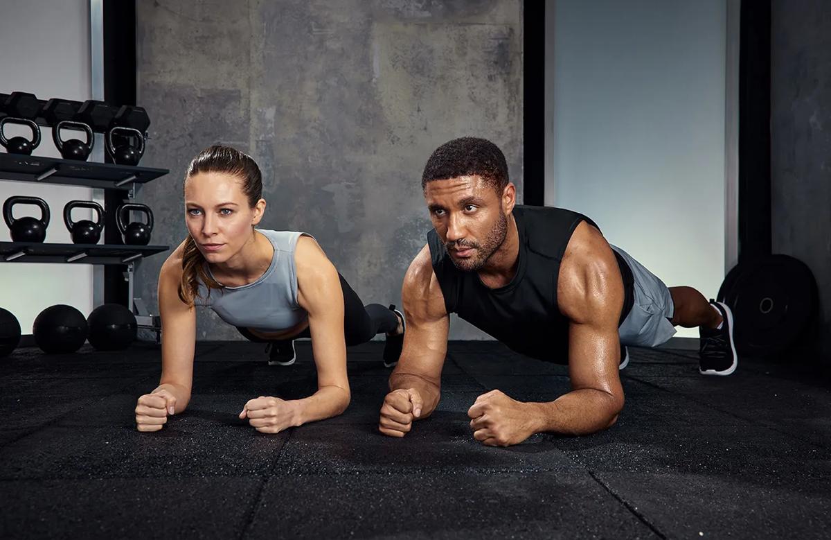 Due persone sul terreno della palestra che mantengono la posizione plank guardando in avanti con i kettlebell sullo sfondo.