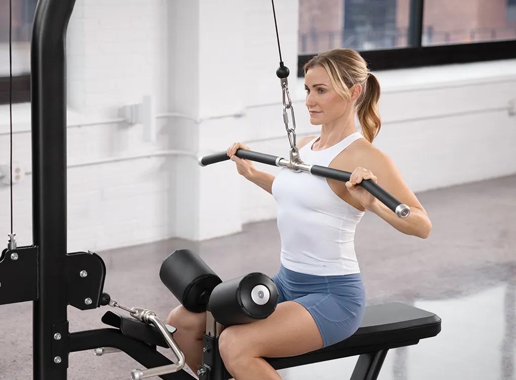 Person using the Keystone Lat Pulldown / Seated Row machine in a lat pulldown position, seated with knees secured under the pad and pulling the bar toward the chest.