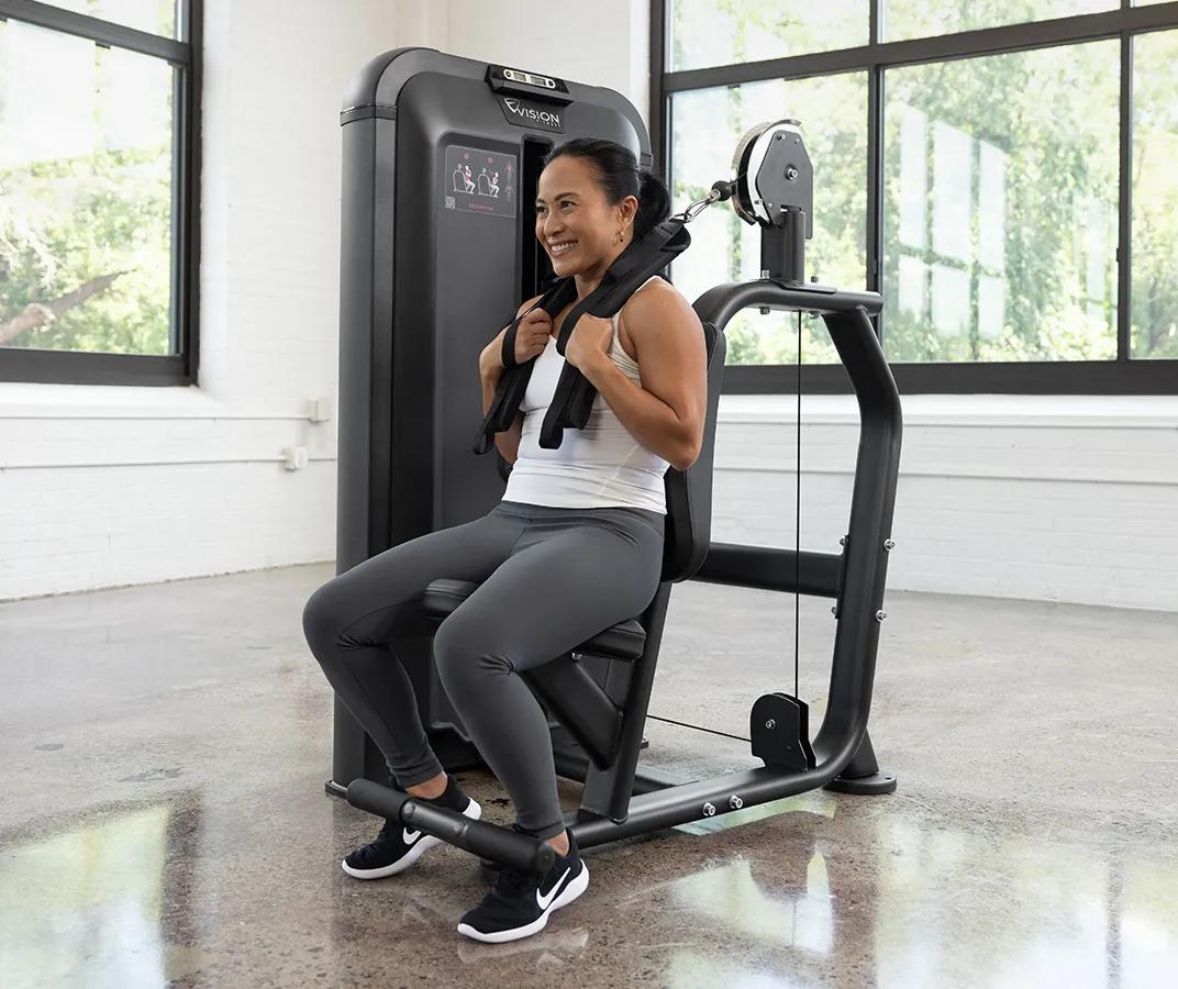 Person seated on the Vision Fitness Abdominal machine with a shoulder harness in place, preparing to perform a crunch in a bright gym setting.