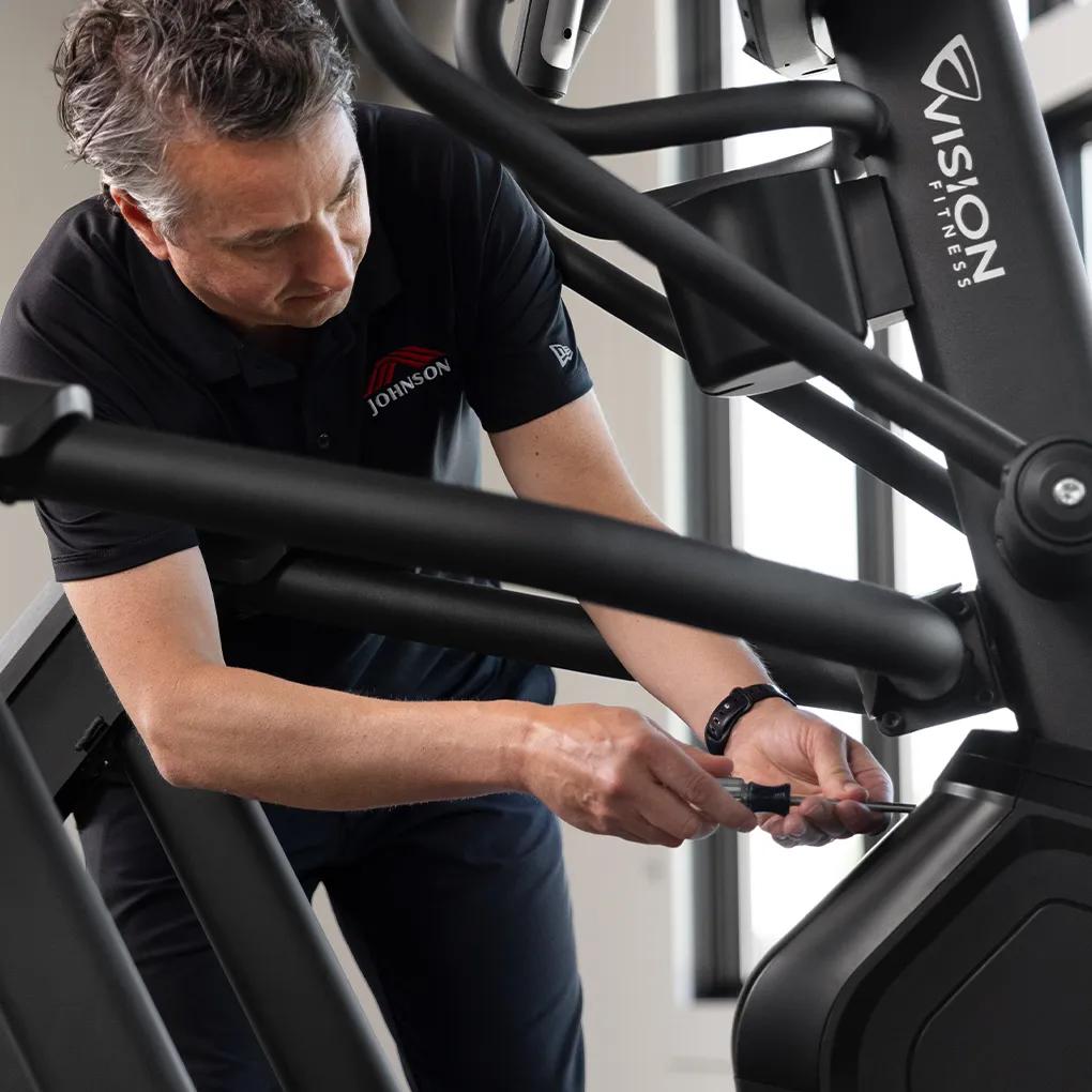A person wearing a Johnson Health Tech shirt using a screwdriver to open the Summit Suspension Elliptical for maintenance.