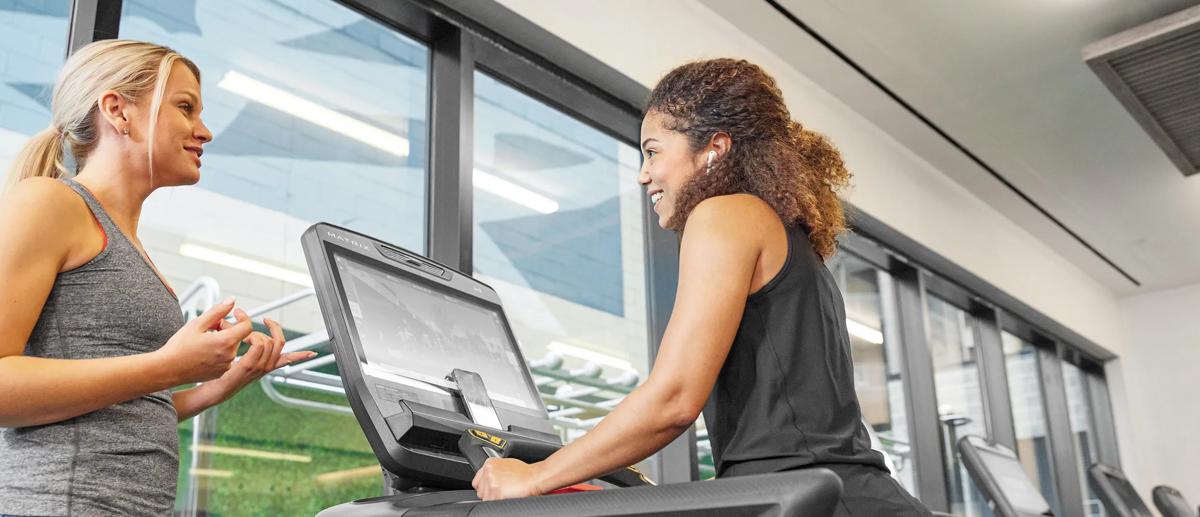 Due persone in abbigliamento sportivo parlano in palestra mentre un'altra è sul treadmill Matrix.